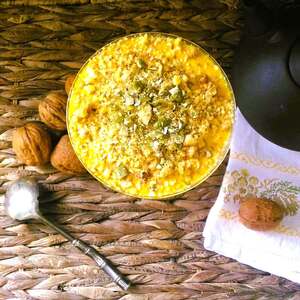 Una foto vista dall'alto che mostra una crema di zucca e ricotta, di colore arancione chiaro, ricoperta da granella di noci e semi di zucca macinati, di colore marrone chiaro e verde. La crema è contenuta in una ciotola rotonda di vetro trasparente. Si vedono una parte di una teiera di colore nero, un canovaccio bianco, noci intere e un vecchio cucchiaio da marmellata, poggiati su una base di paglia intrecciata.